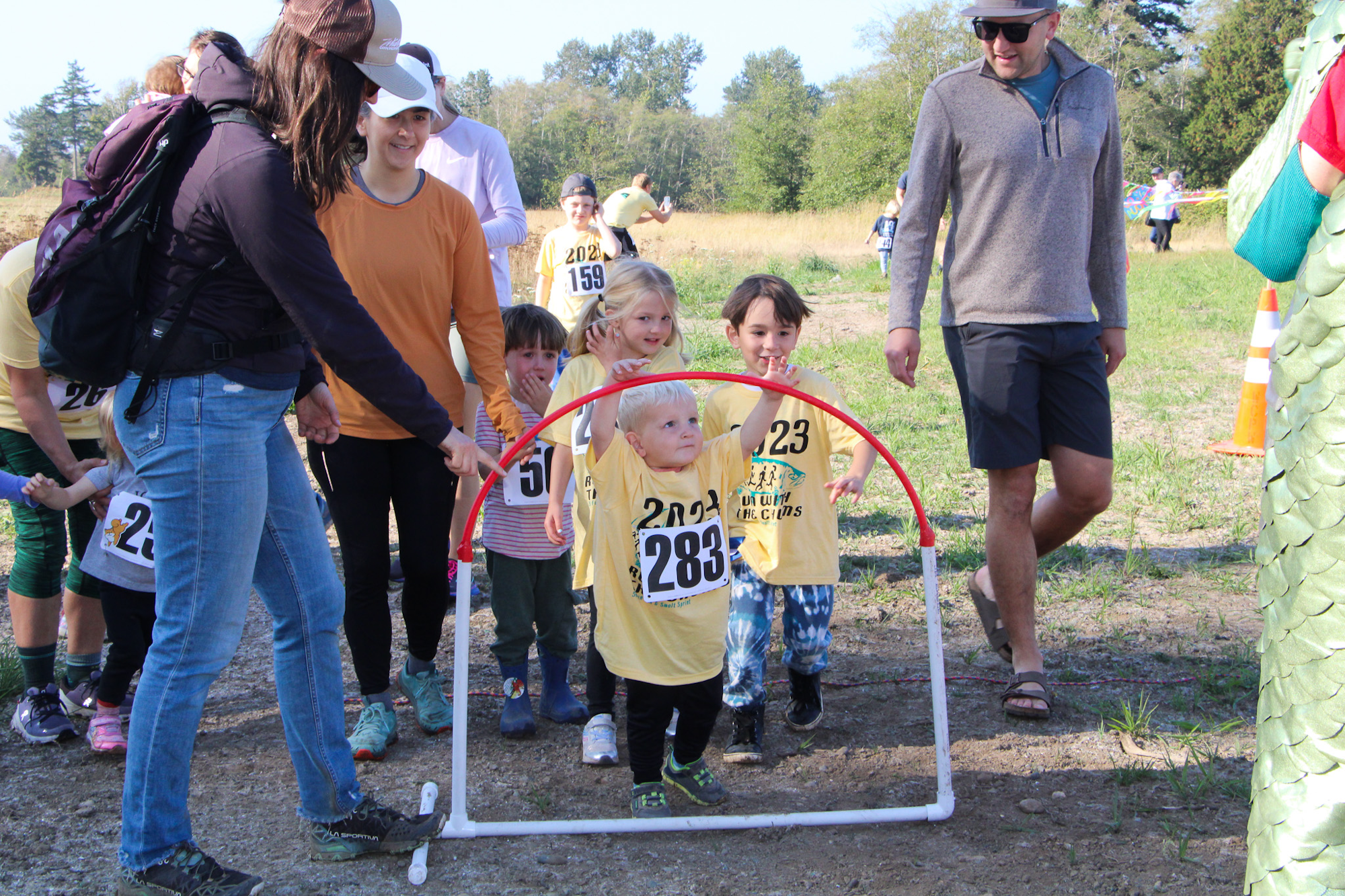 Children with race numbers gather at an outdoor event, surrounded by adults and a small hoop.