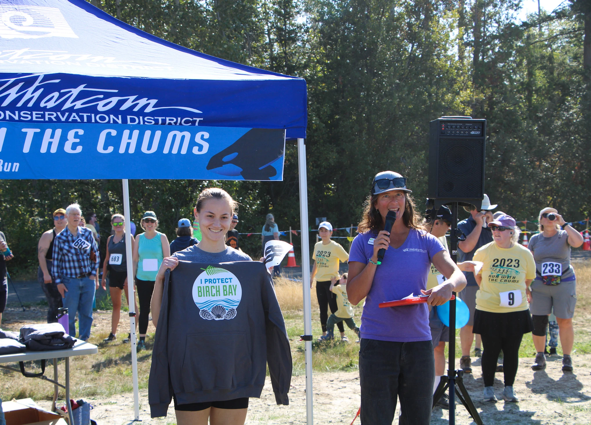 Outdoor event with runners, a woman holding a "I Protect Birch Bay" shirt, and a speaker under a "Run with the Chums" tent.