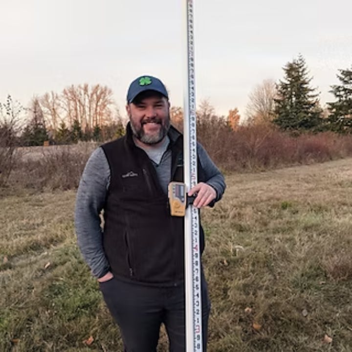 A person standing outdoors holding a measuring rod in a grassy area.