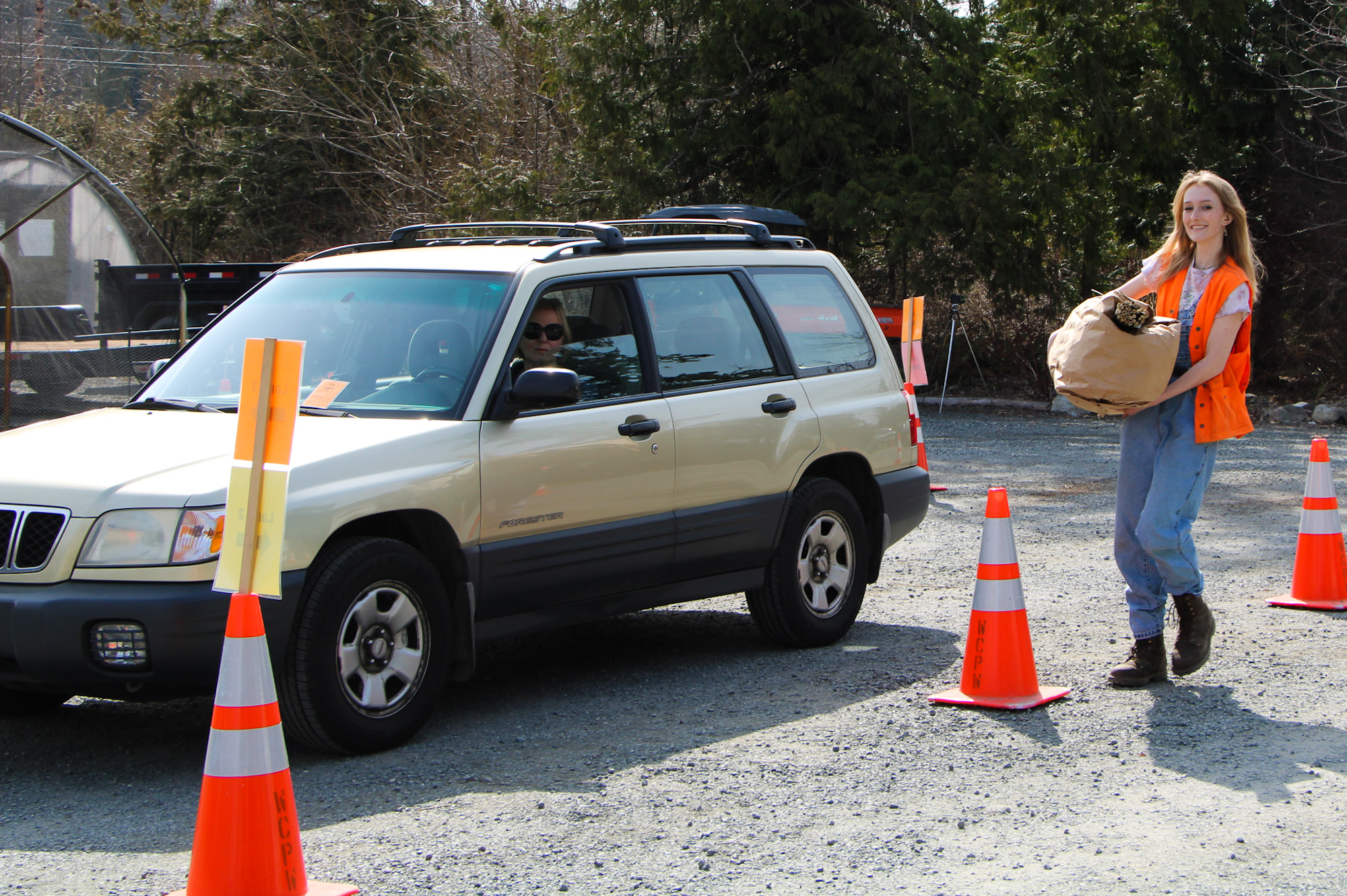 A woman in an orange vest carries a bag. A car is parked nearby, with traffic cones along the road.