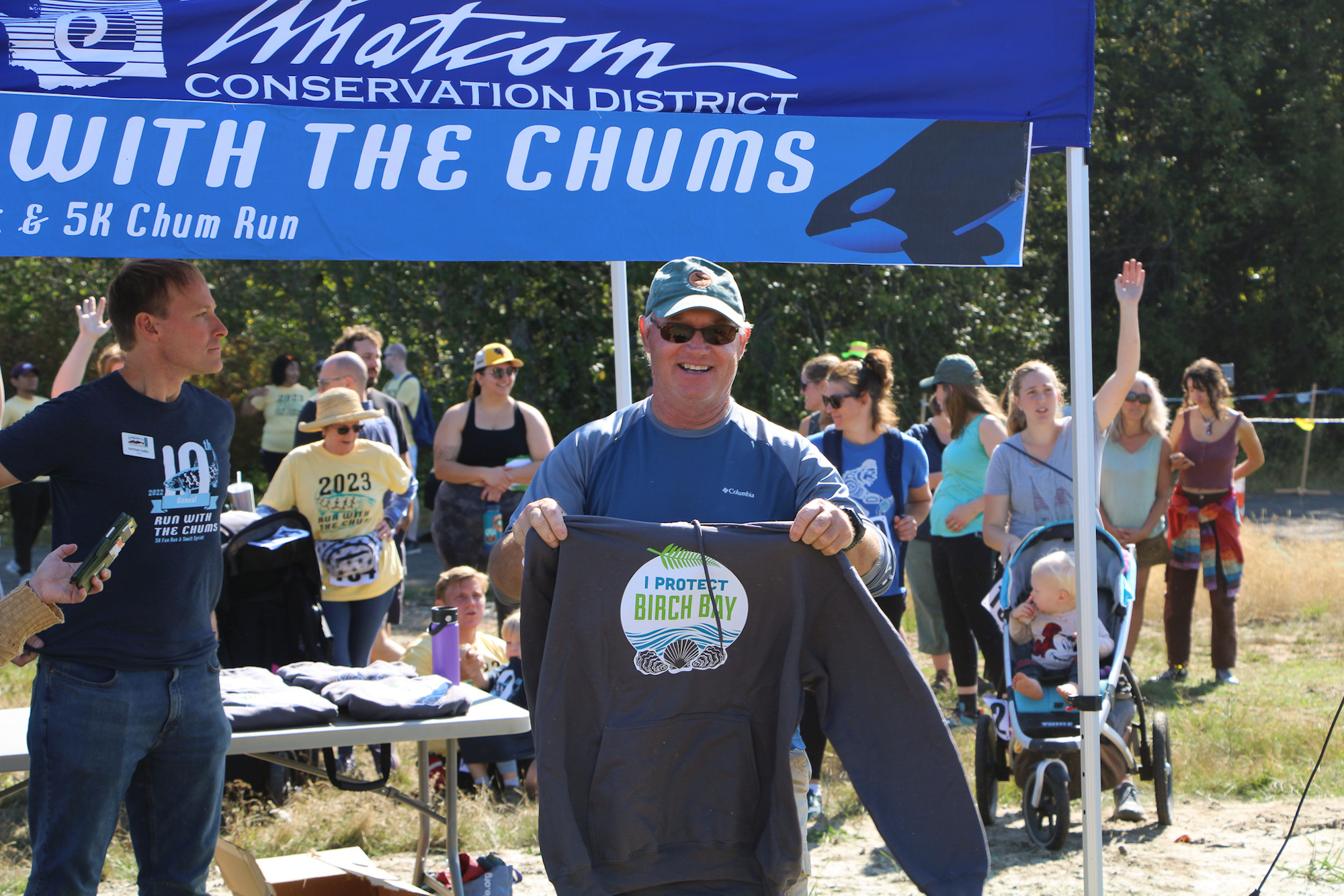 Outdoor event with a man showing a "Protect Birch Bay" shirt, people gathered under a tent for "Run with the Chums" by Whatcom Conservation.