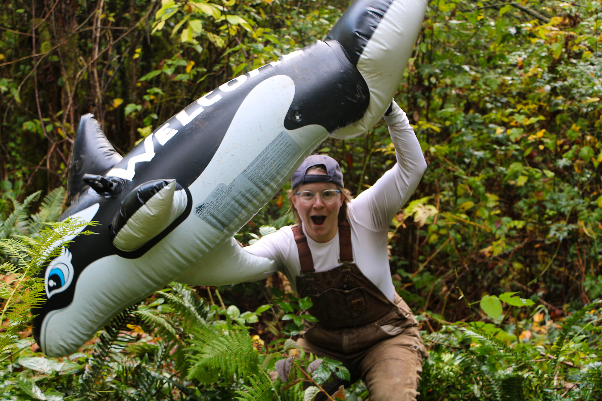 A person in overalls, excitedly holding a large inflatable orca in a forest.