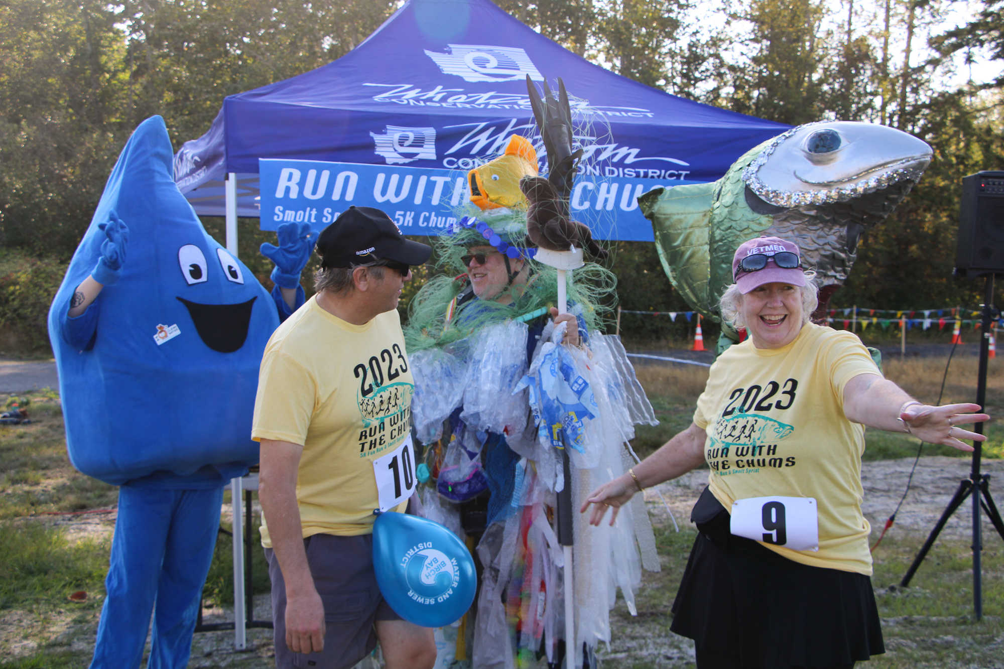 People in costume and race attire at a "Run with the Chums" event, featuring a blue mascot and a fish sculpture.