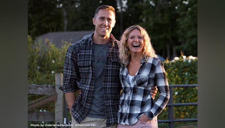 A man and woman smiling, standing outdoors, both wearing plaid shirts.