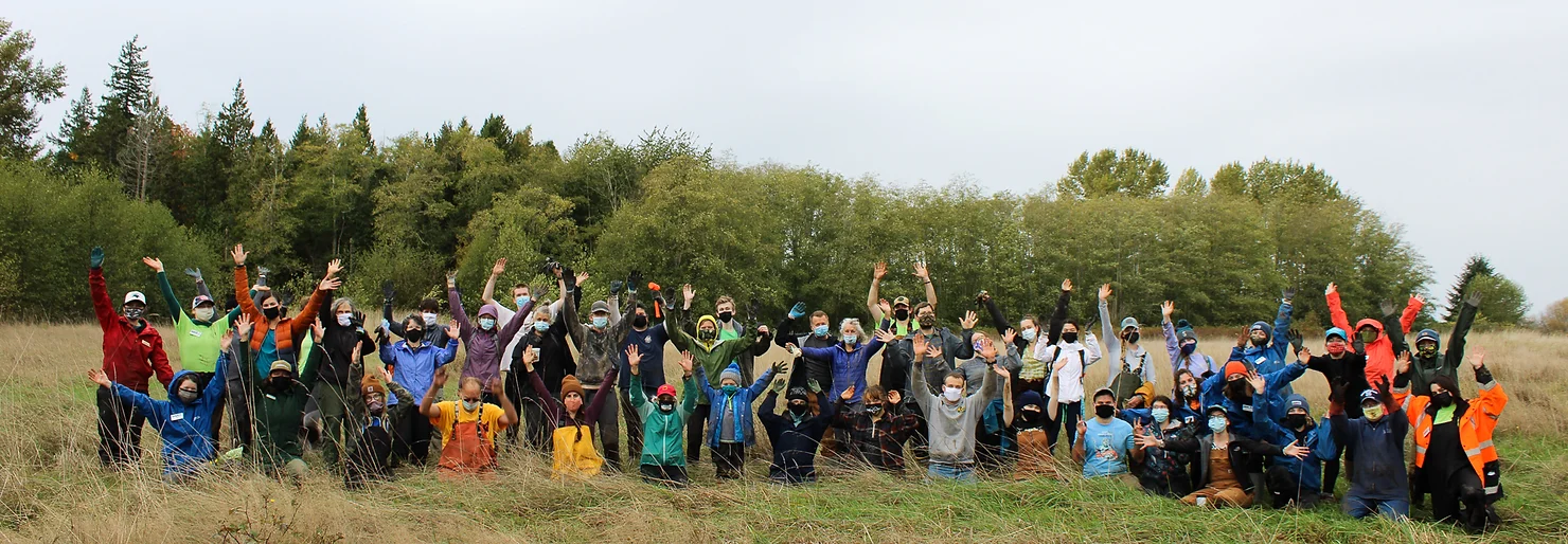 A group of people outdoors, wearing masks, waving their hands in a field with trees in the background.