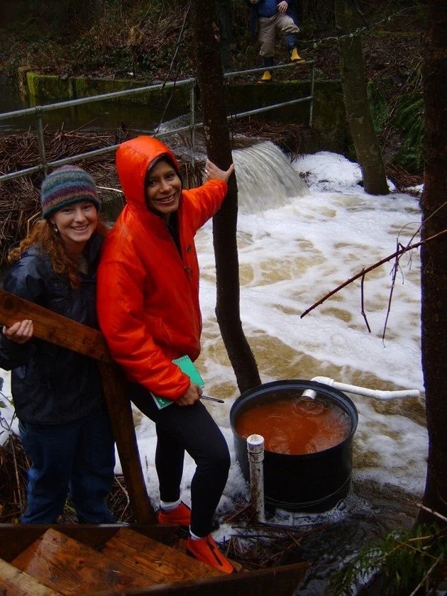 Two people in rain gear stand by a flowing stream with a water sample collection barrel nearby.