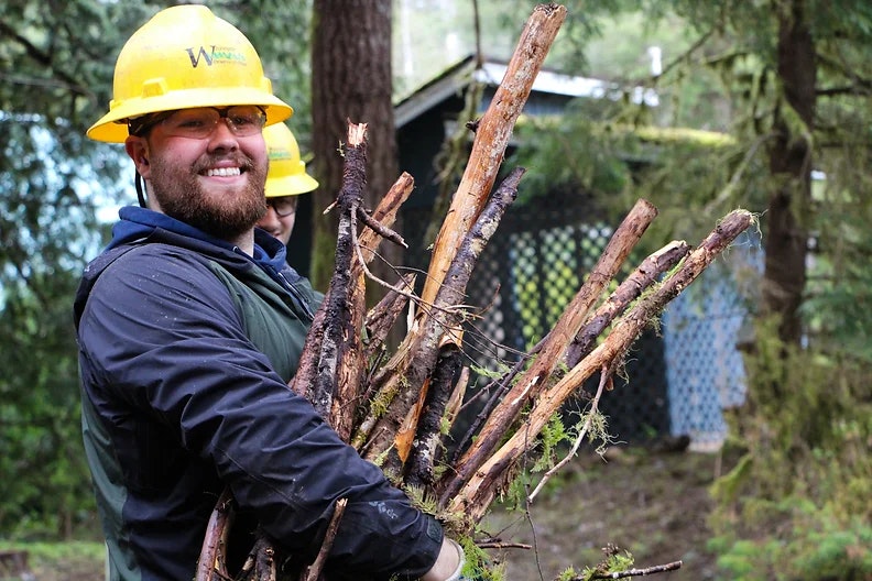 A person wearing a yellow hard hat and glasses, smiling while holding wooden branches in a forest setting.