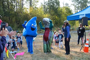 Costumed figures and children at an outdoor event, with spectators and a speaker on stage.