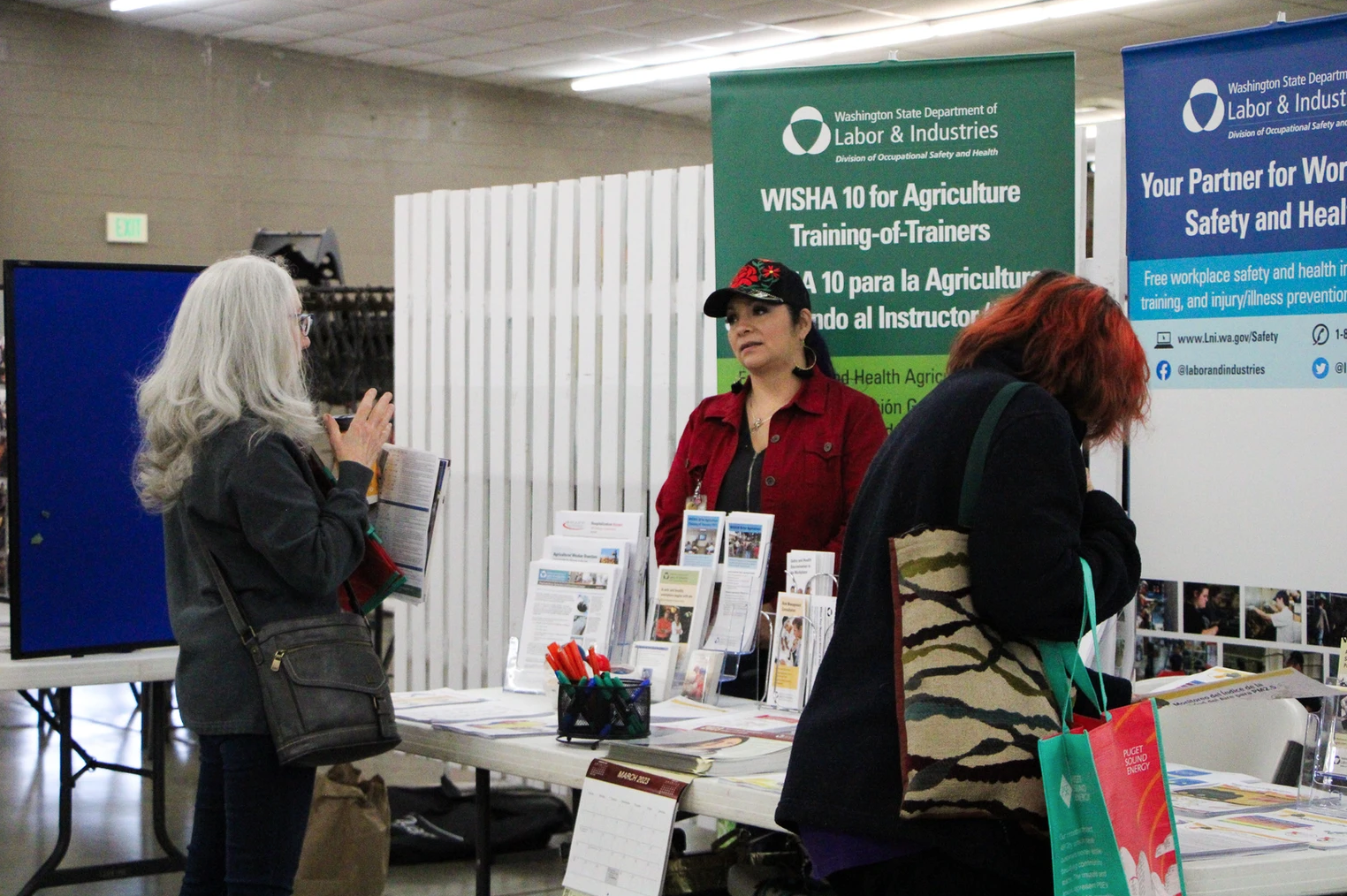 Three people at a Washington State Labor & Industries booth with brochures and banners about agriculture training and workplace safety.