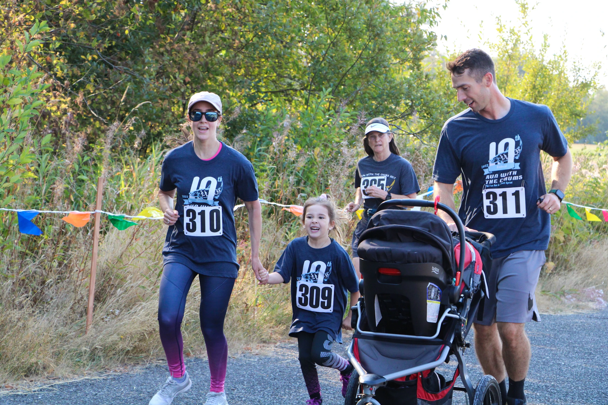 A group of people, including a child, running in a race, some with race numbers, and one pushing a stroller, on a tree-lined path.