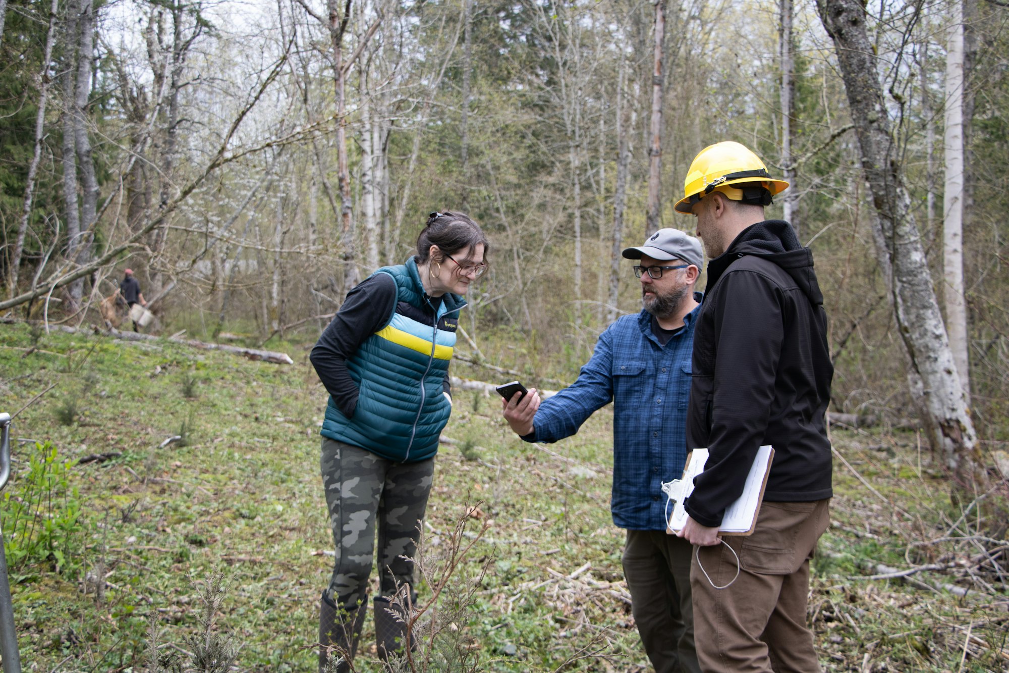 A group of people in a forest discussing something on a phone, with one wearing a hard hat and another holding papers.
