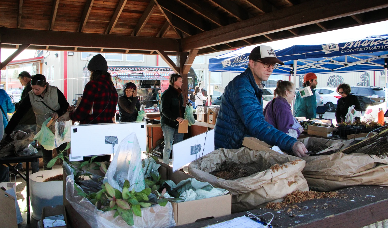 People at an outdoor market under a wooden shelter, handling plants and packaged items.