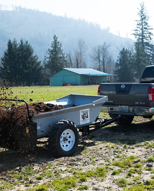 A truck with a trailer spreading soil or compost on a grassy field with trees and a building in the background.