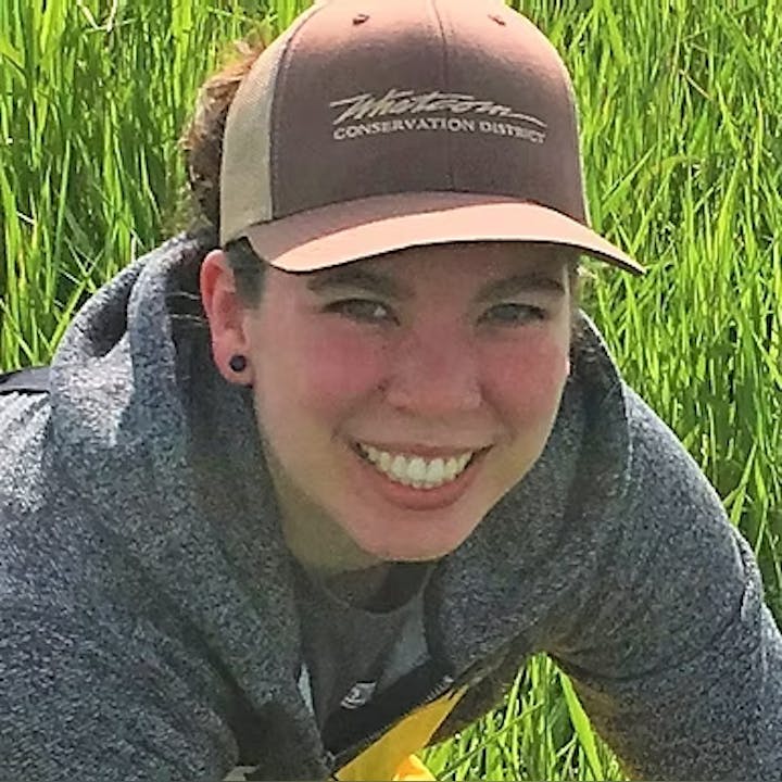 A person smiling, wearing a cap and hoodie, in a grassy area.
