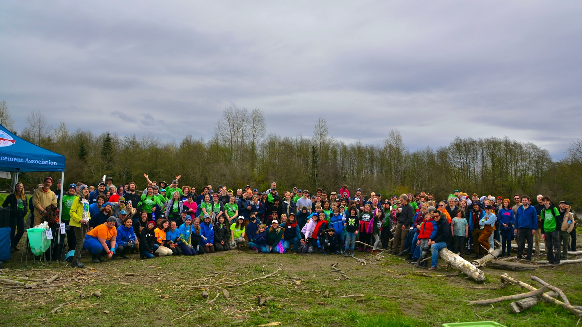 A large group of people posing outdoors near a tent, with trees in the background.