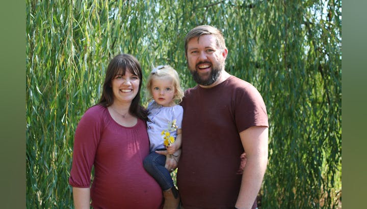 A smiling family of three stands in front of lush greenery, holding yellow flowers.