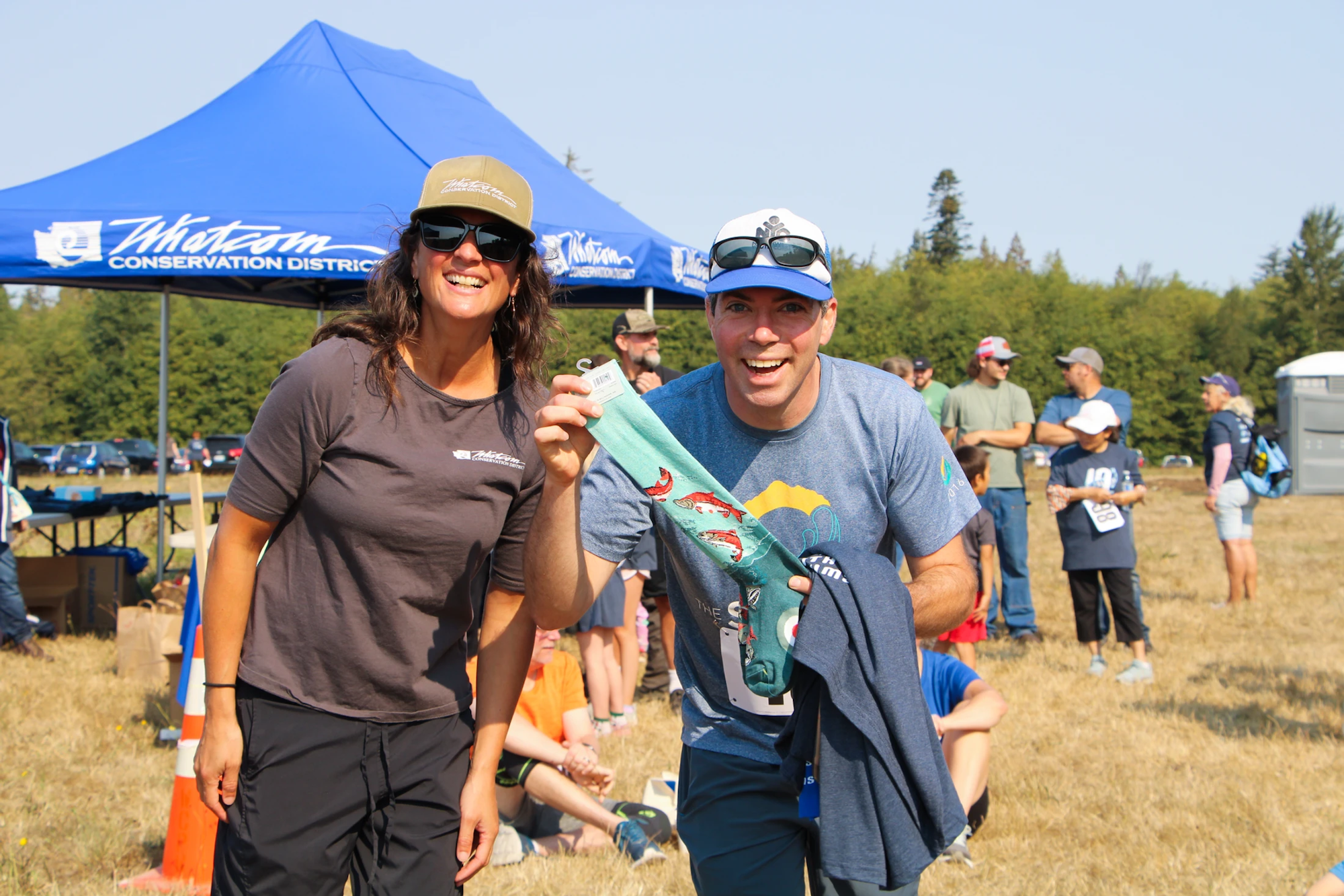 Two people smiling outdoors with a colorful sock, in front of a "Whatcom Conservation District" booth.