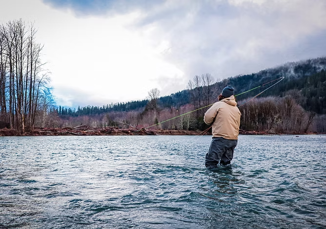 Person fly fishing in a river surrounded by trees and hills under a cloudy sky.