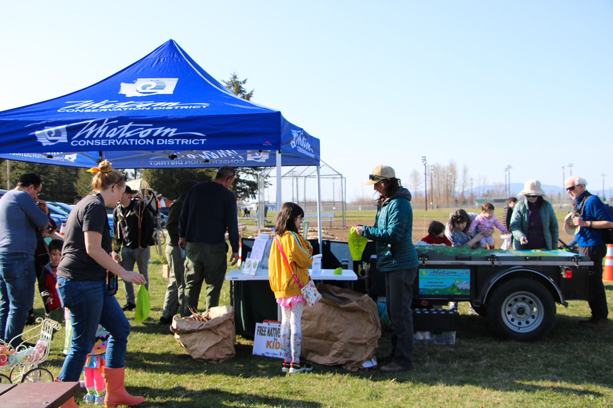 Outdoor event with people gathered under a blue "Whatcom Conservation District" tent, interacting and distributing items.