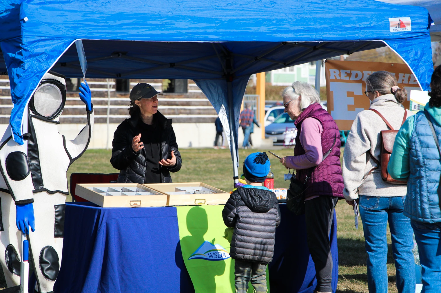 People gathered under a blue tent with a table, a mascot in a suit, and signs.