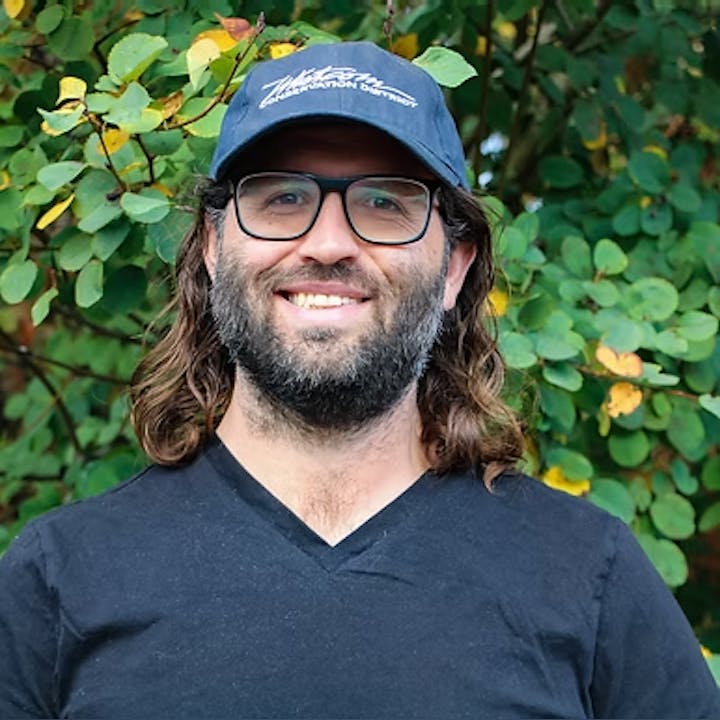 A person with glasses and a cap, wearing a black shirt, stands in front of green foliage, smiling.