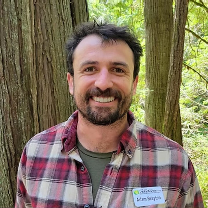 A person with a beard wearing a plaid shirt, smiling in a forest setting.