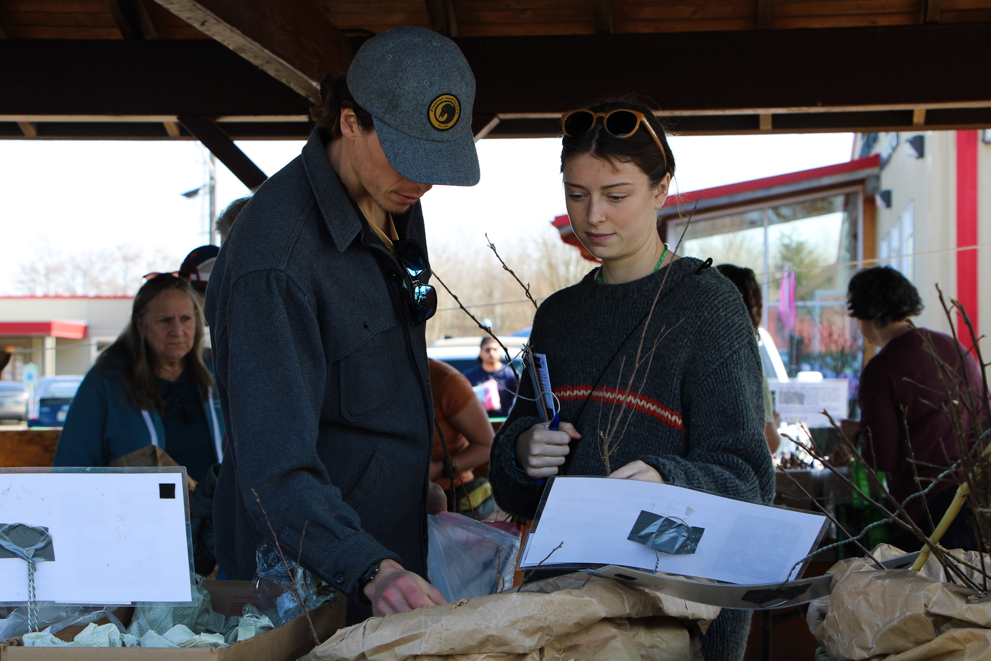 Two people examining plants at an outdoor market with others in the background.