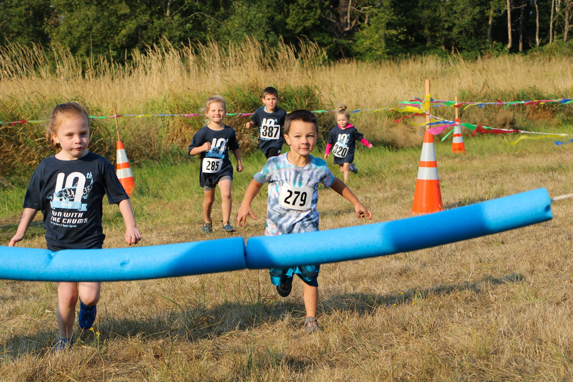 Children running a race in a grassy field, wearing numbered shirts and crossing a pool noodle finish line.