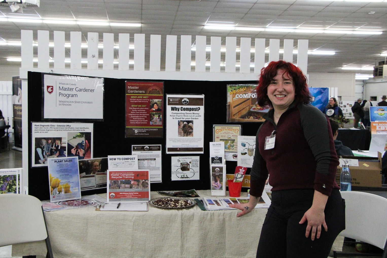 A person standing by a Master Gardener Program display with composting info, smiling at an indoor event.