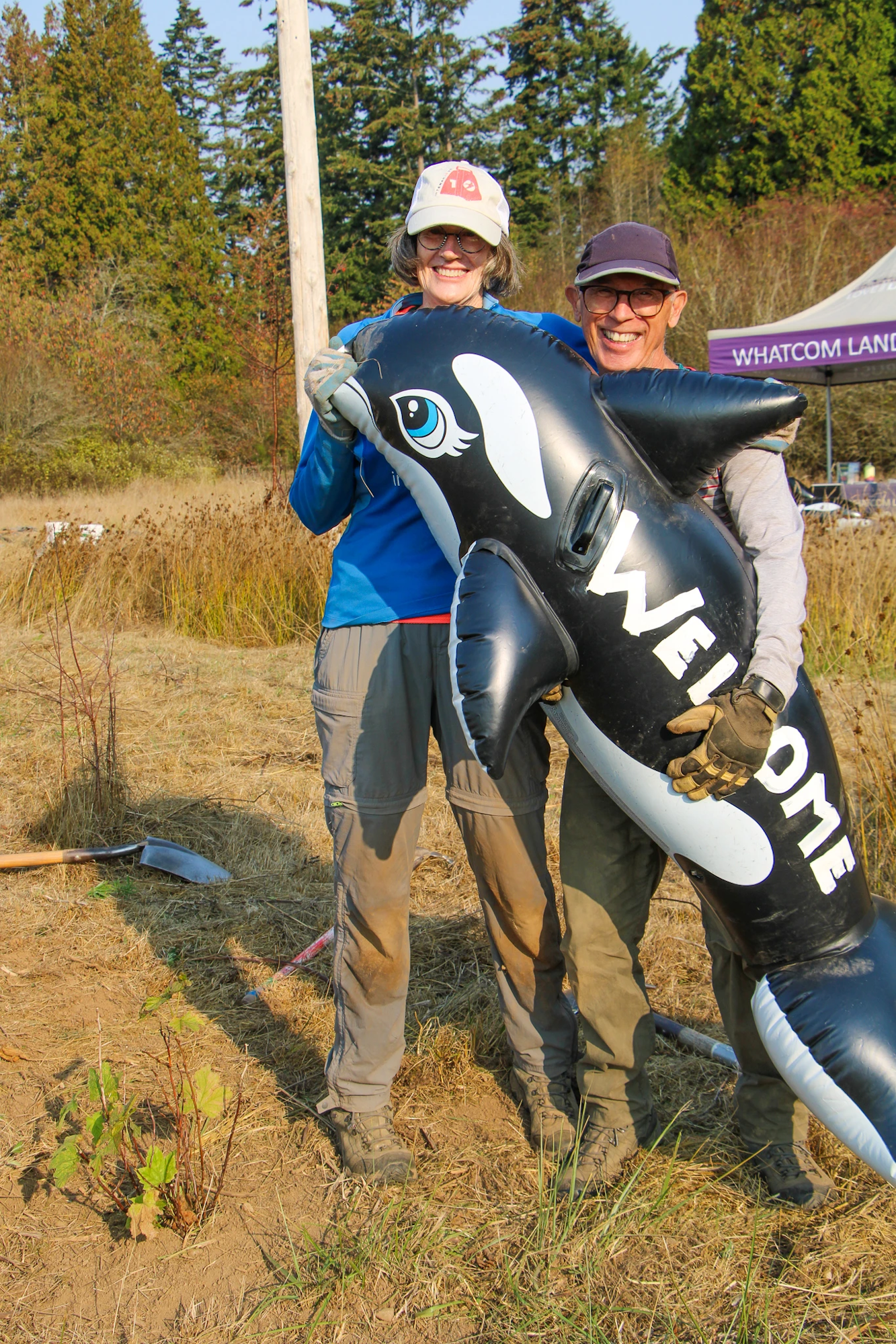 Two people outdoors holding a large inflatable orca with "welcome" written on it.