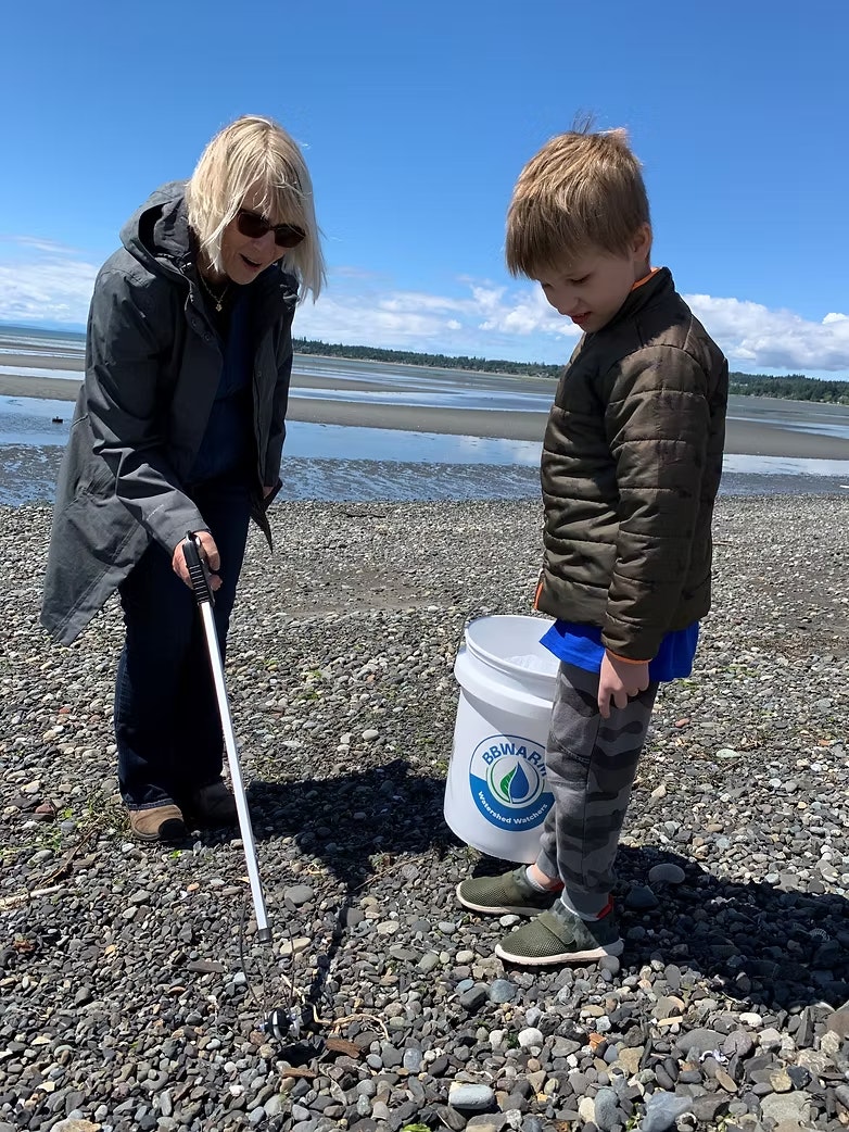 A woman and a child are cleaning a rocky shore, one holding a pick-up tool and the other a bucket.