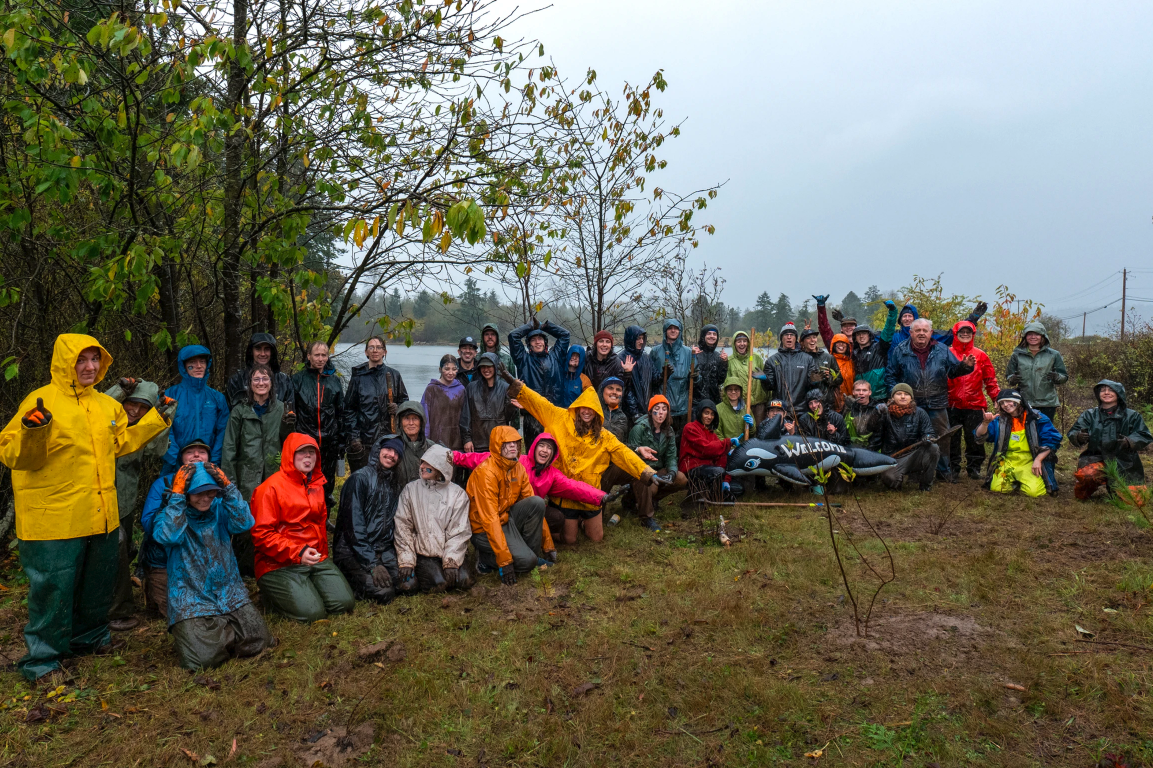 A group of people in raincoats gather outdoors near trees and a lake, some holding an inflatable dolphin labeled "WELCOME."