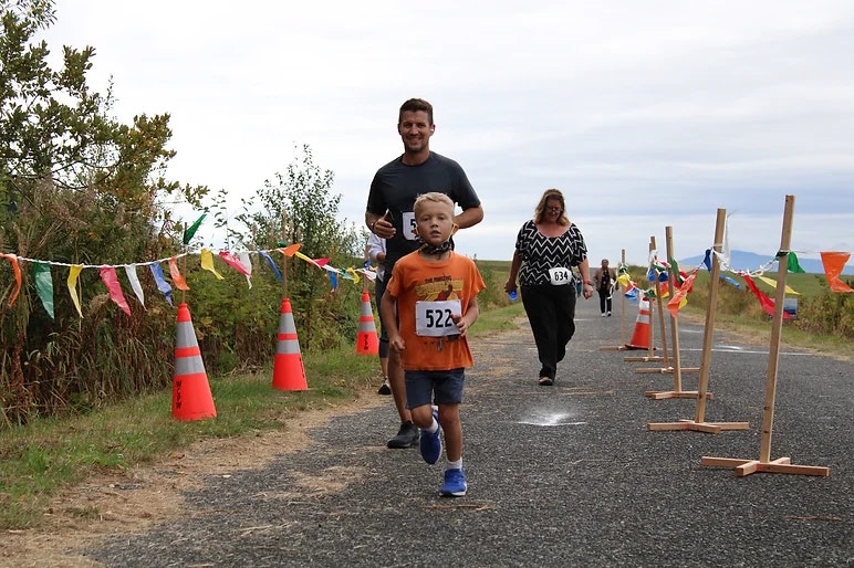 A child in an orange shirt and blue sneakers runs a race, followed by two adults, with colorful flags and cones along the path.