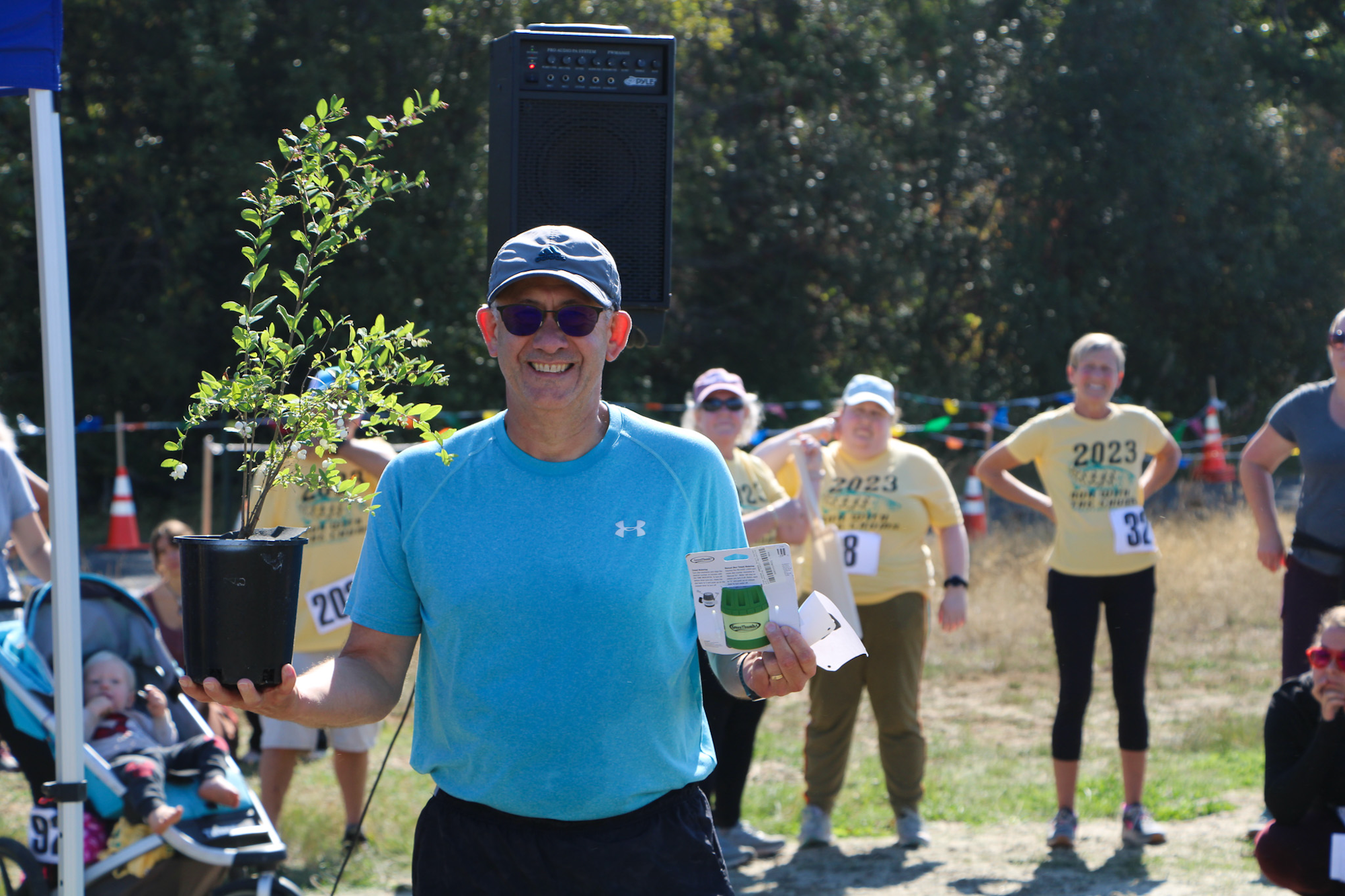 A man in a blue shirt holds a potted plant and a card. People in yellow shirts stand in the background with cones and a stroller nearby.