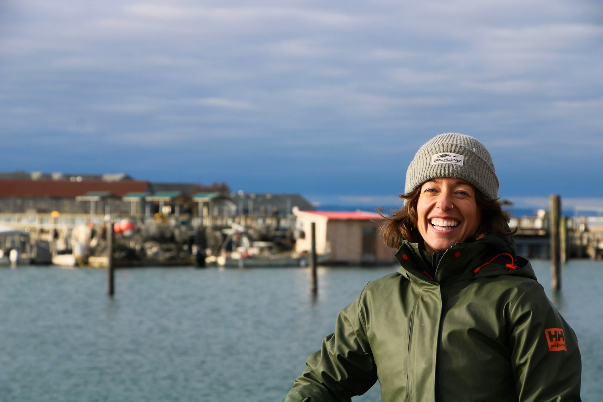 A person in a green jacket and gray beanie smiles by the water with a dock and buildings in the background.