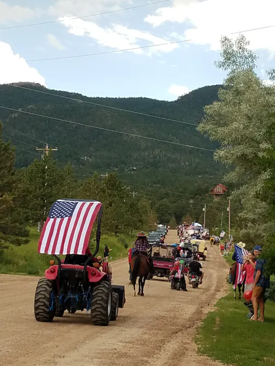 A festive parade featuring tractors, horses, and vehicles decorated with American flags on a dirt road in a scenic area.