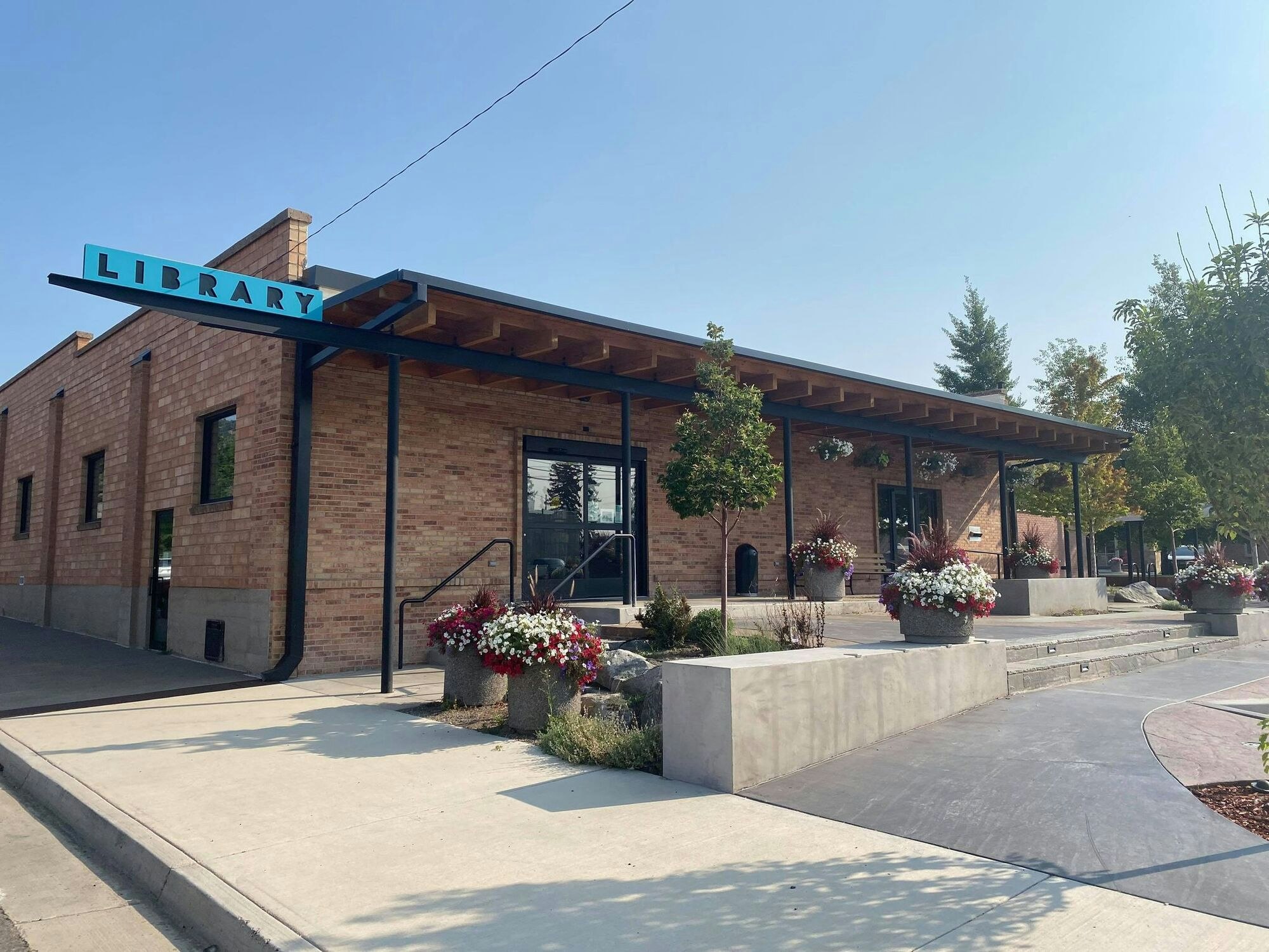 A modern library building with a brick exterior, colorful flower pots, and a clear blue sky above.