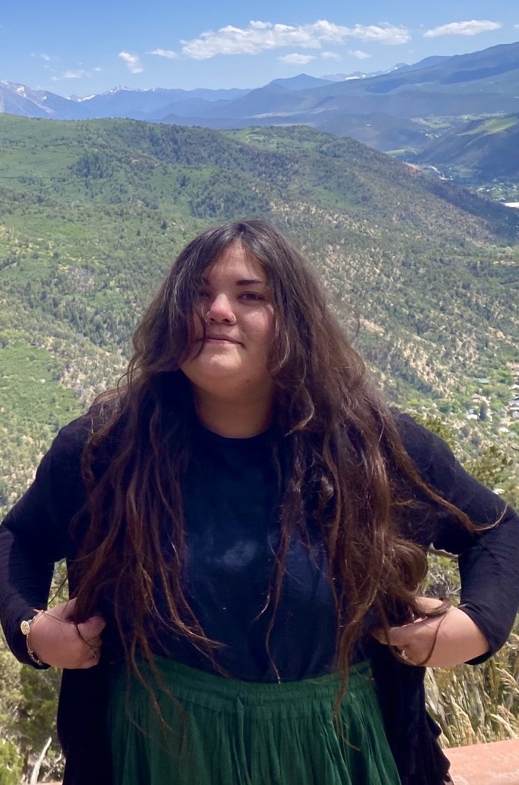 A person with long hair stands outdoors, smiling against a backdrop of mountains and greenery. Bright, sunny day.