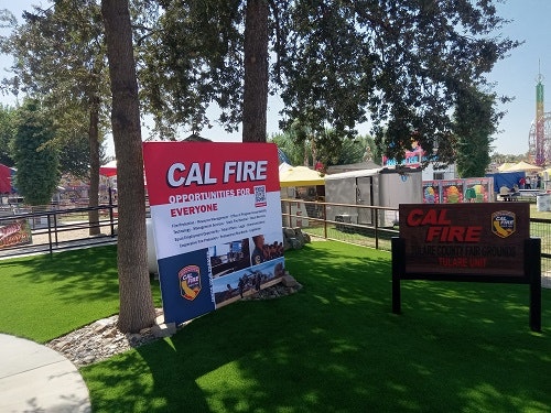 A CAL FIRE recruitment sign stands in a park with trees, artificial grass, and amusement park rides in the background.