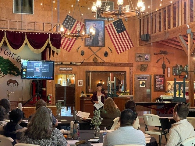 A rustic room with people seated, an American theme, flags, a person speaking at a podium, wooden interior, and loft area.