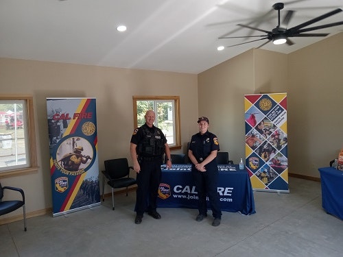 Two uniformed individuals stand by CAL FIRE recruitment banners and a table.