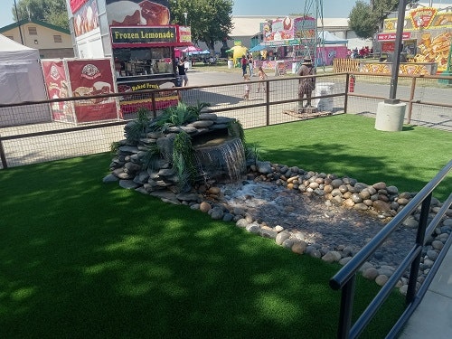 Small artificial waterfall on artificial grass with food stands in the background at an outdoor event.