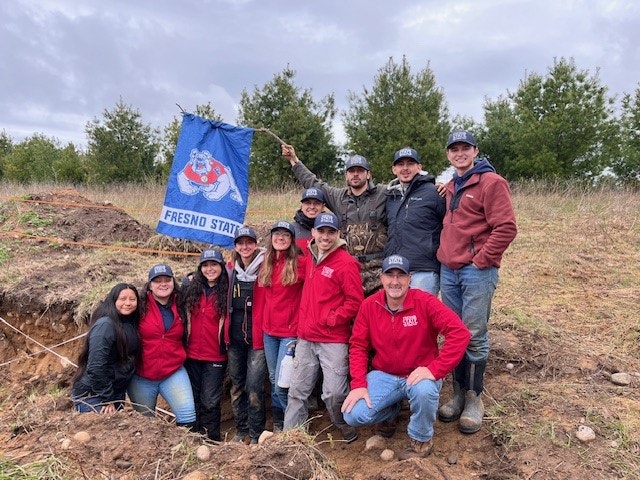 A group of people outdoors, some kneeling in a trench, holding a Fresno State flag, wearing red and dark jackets.