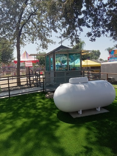 A small building surrounded by trees, tents, and a white propane tank on artificial grass.
