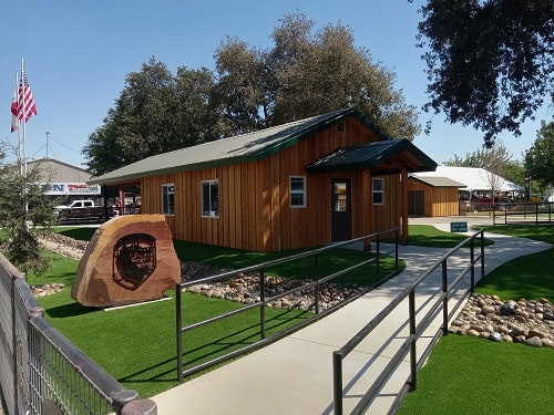 A small wooden building with a flag, ramp, and a stone sign in front, surrounded by trees and artificial grass.