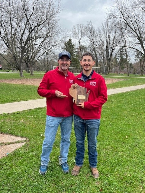 Two people in red jackets, one holding a wooden plaque, stand on grass with bare trees in the background.