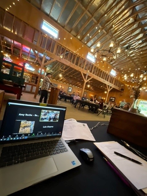 A laptop on a table with a presentation, a person standing at a podium, and attendees seated at tables in a room with a wooden ceiling and lights.