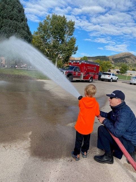 A child with an adult holding a fire hose, spraying water, with a fire truck in the background.
