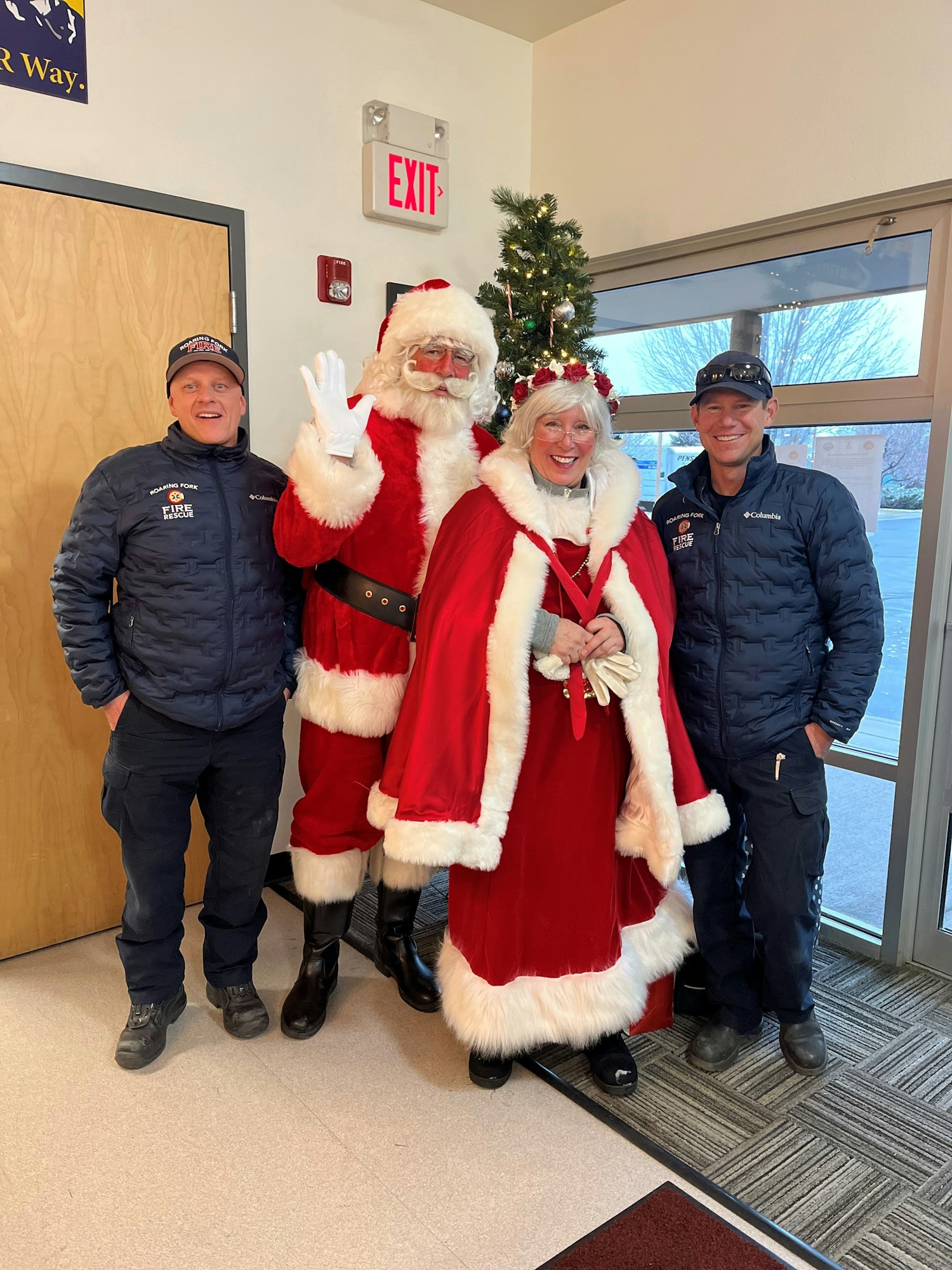 Santa Claus and Mrs. Claus posing with two firefighters by a Christmas tree indoors.