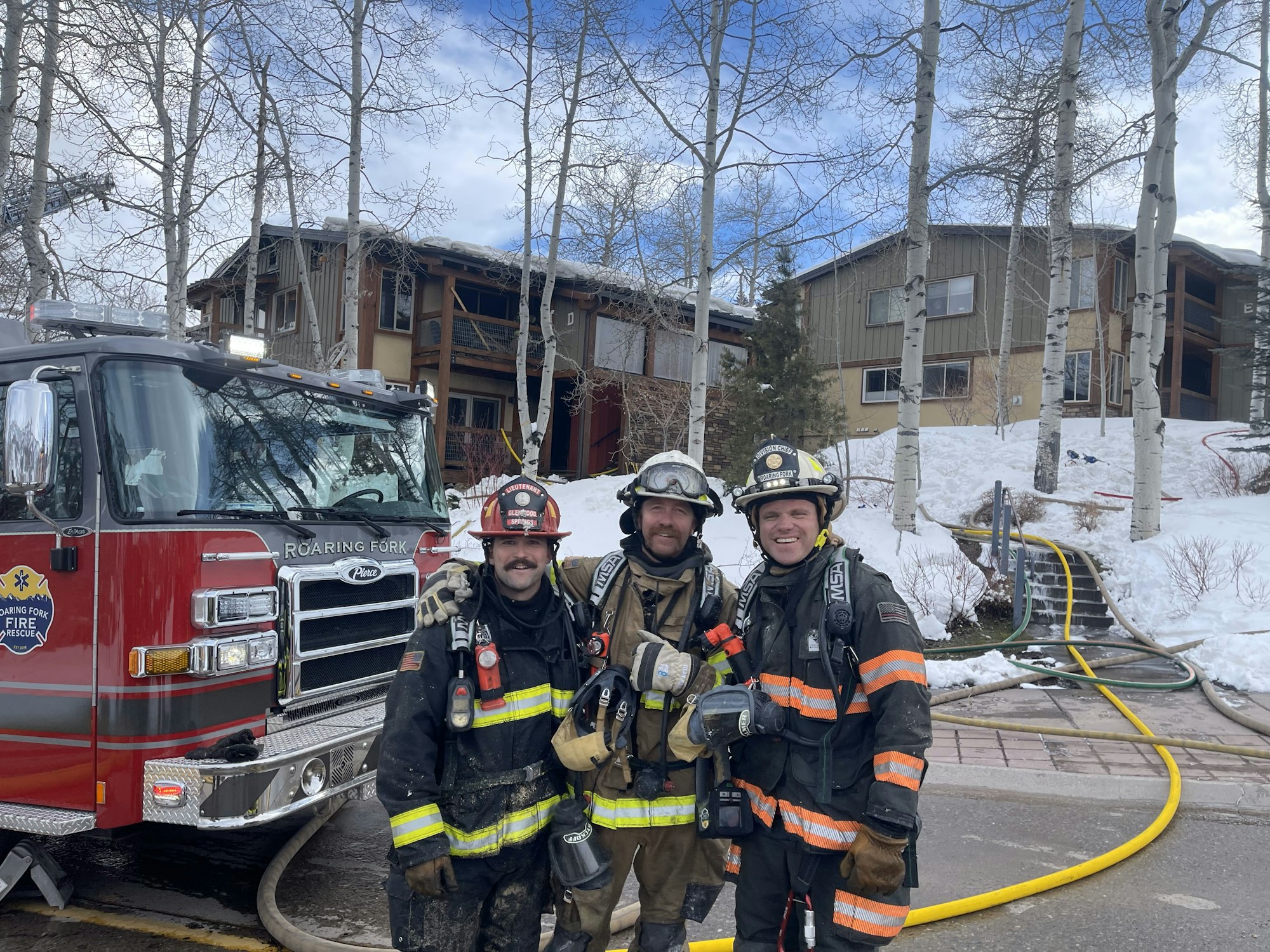 Three firefighters in front of a fire truck, damaged building in the background, with snow around.